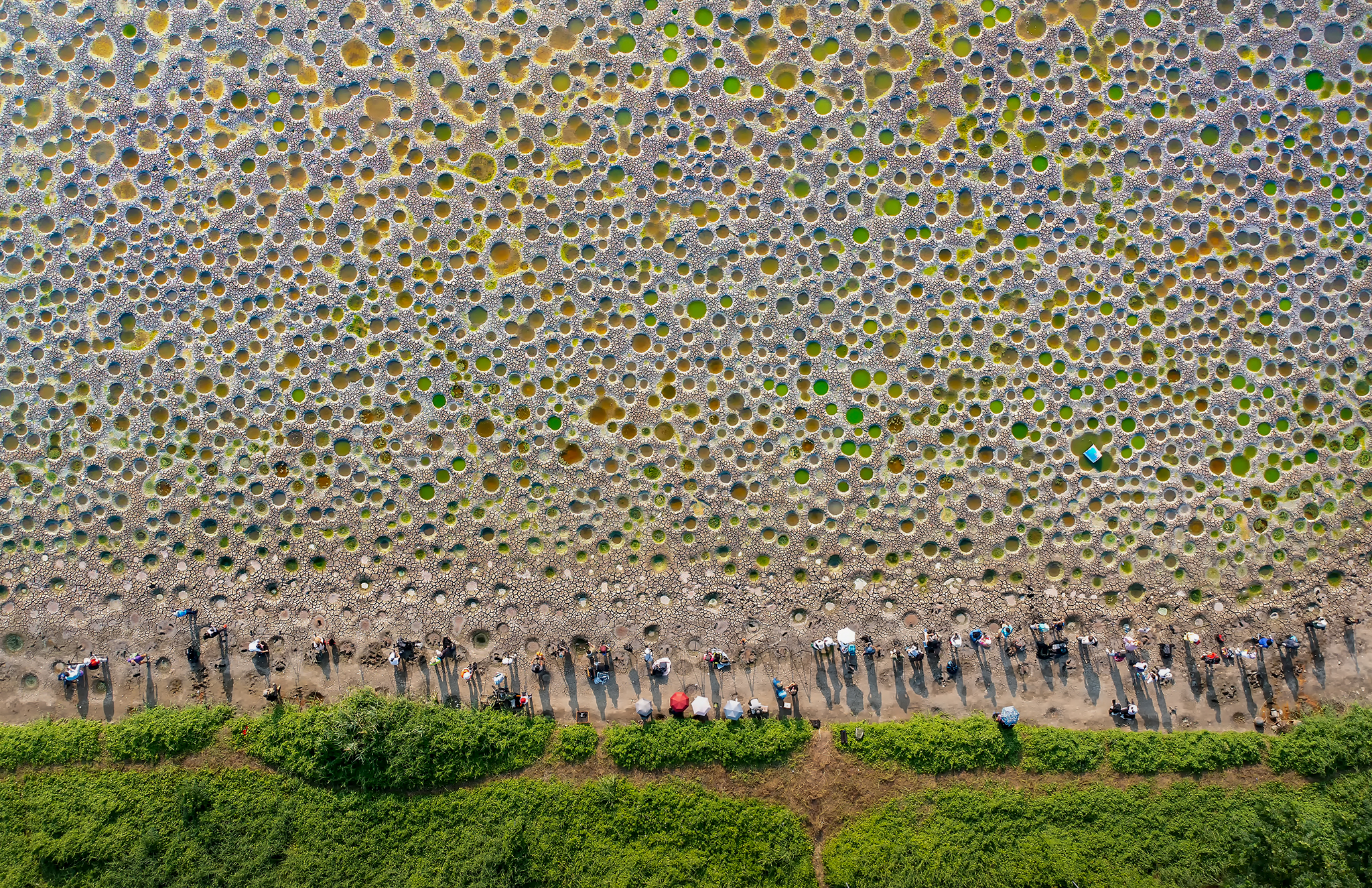 A group of people stand at the edge of a large, open sandy area. Large holes dot the surface, filled with green and orange water.